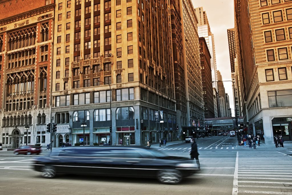 Busy downtown street scene with tall historic and modern buildings, pedestrians crossing, and a black limousine driving by.