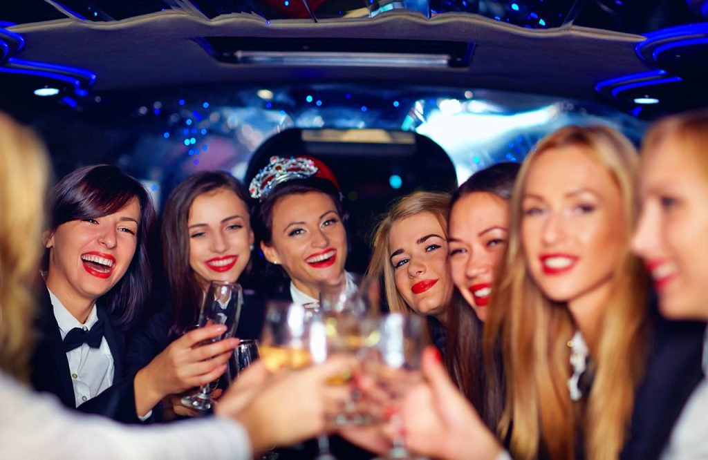 Group of smiling women dressed for a party, raising glasses in a toast inside a limousine with colorful interior lighting.