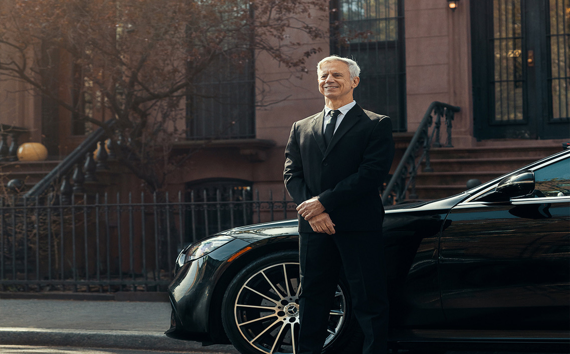 Chauffeur in a black suit standing beside a luxury black sedan, with classic brownstone buildings in the background.