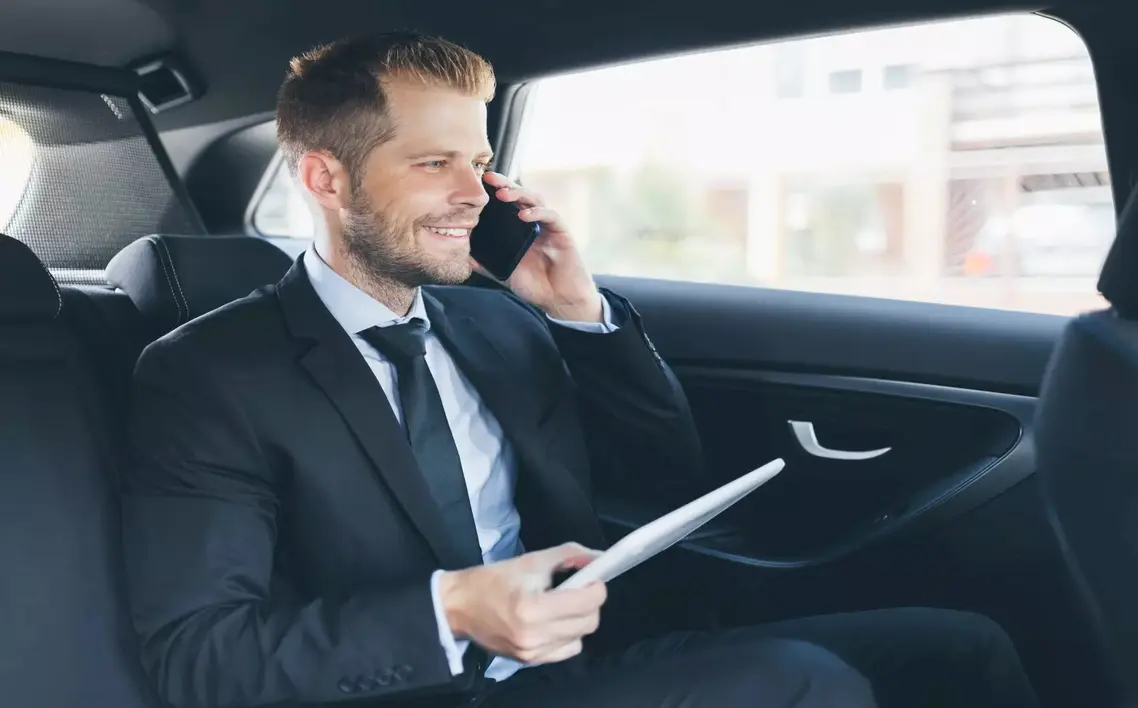 Businessman in a suit sitting in the back seat of a car, smiling while talking on a smartphone and holding a tablet.