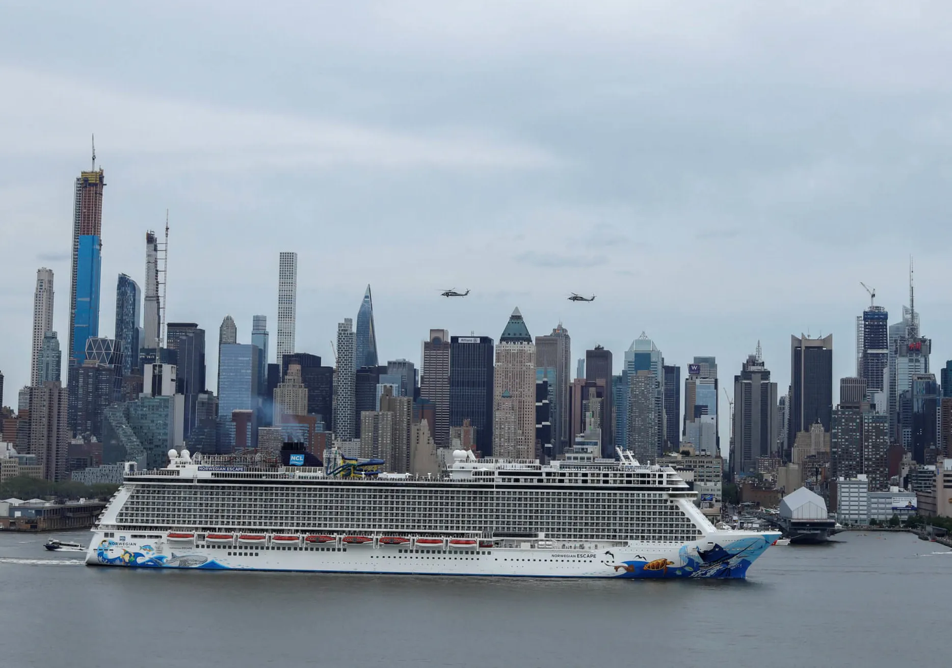 Cruise ship sails past Midtown Manhattan on the Hudson River with helicopters overhead and skyscrapers in view.
