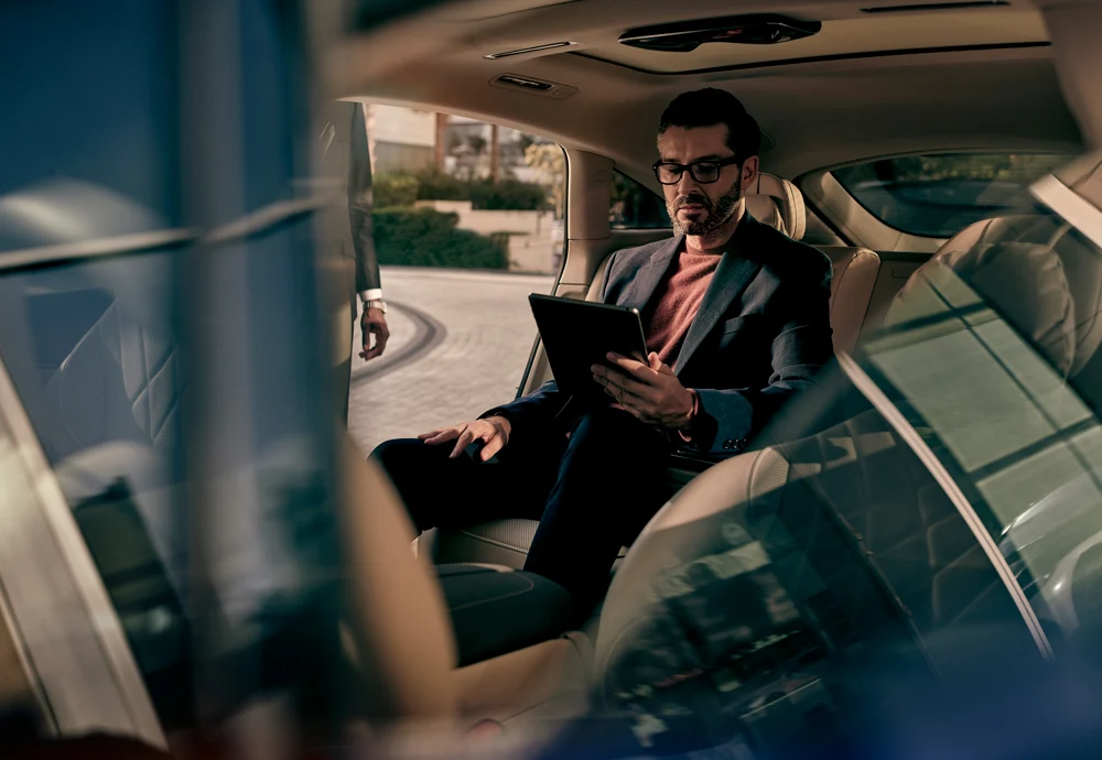 Businessman in the back seat of a luxury car, working on a tablet, with a chauffeur standing outside the vehicle.