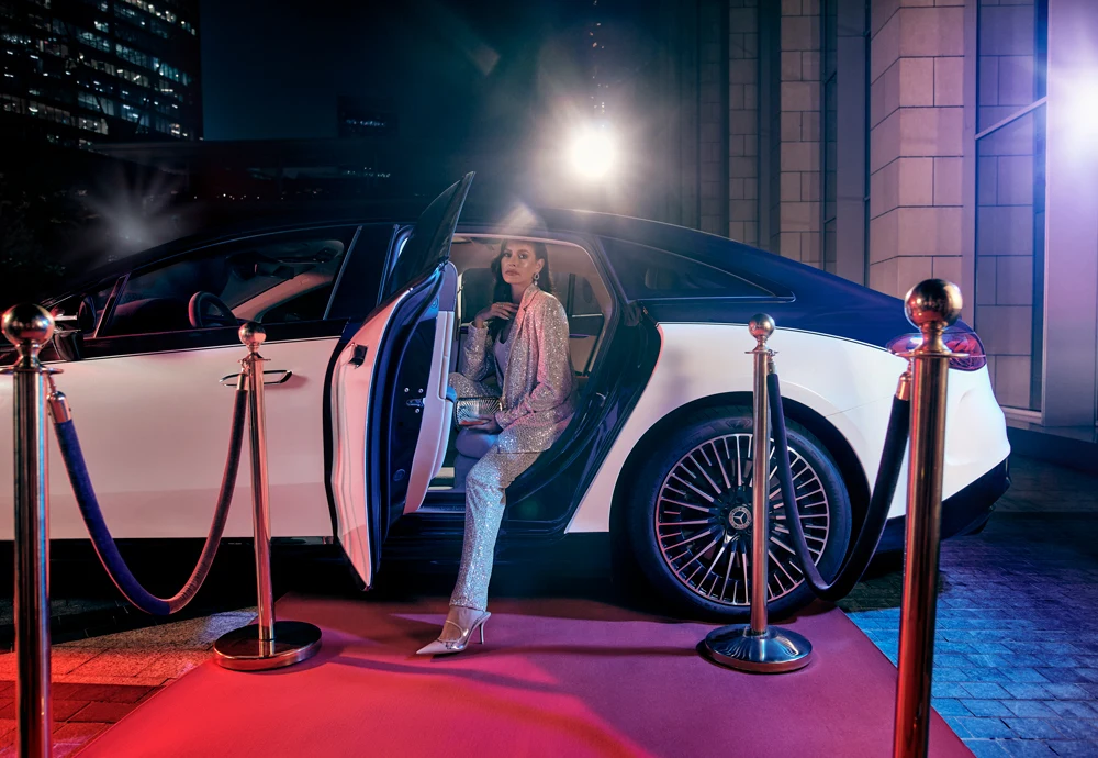 Elegant woman stepping out of a luxury car onto a red carpet, surrounded by velvet ropes and bright event lights.