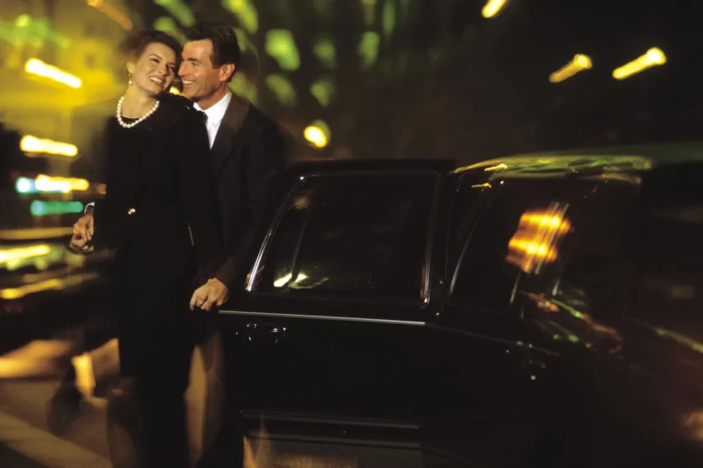 Elegant couple smiling beside a black limousine at night, surrounded by blurred city lights and a romantic atmosphere.