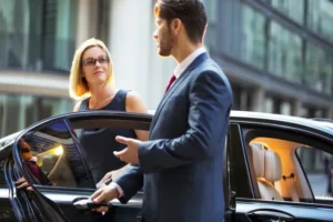Business executive stepping out of a luxury black car with the assistance of a professional chauffeur in a city setting.