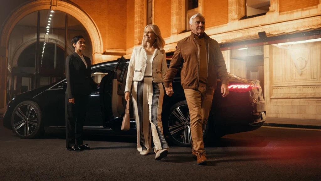 Elegant couple stepping out of a luxury car, greeted by a chauffeur outside a grand, illuminated building entrance.