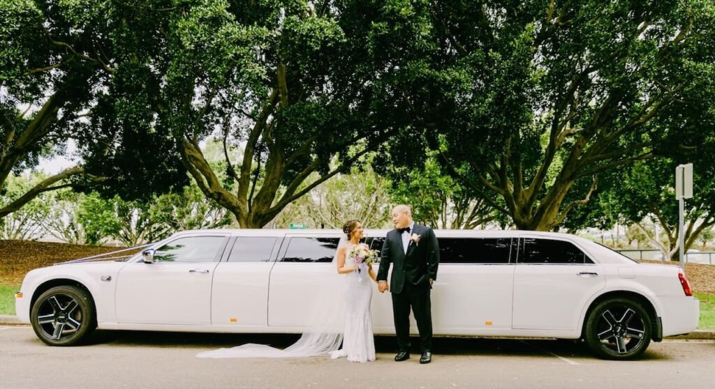 Bride and groom standing in front of a white Chrysler super-stretch limousine under large green trees, ideal for a wedding transport service.