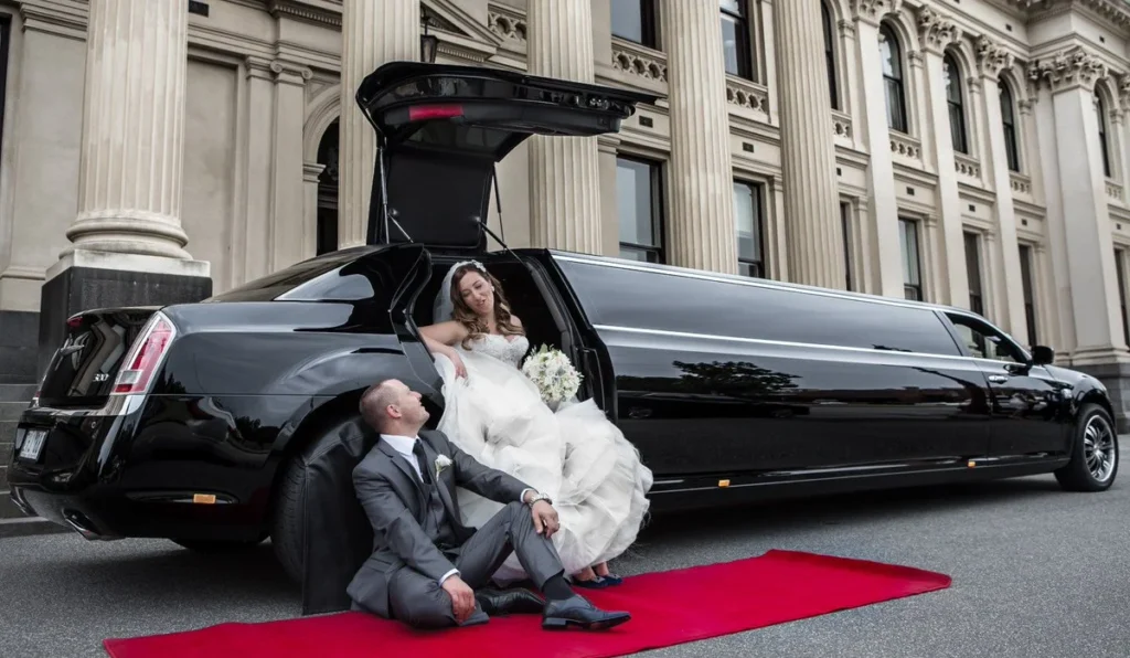 Bride and groom posing beside a luxurious black wedding limousine with red carpet in front of a grand building.