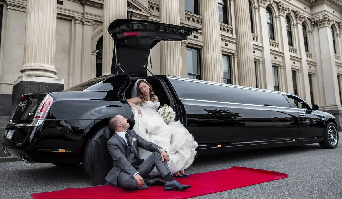 Bride and groom posing beside a luxurious black wedding limousine with red carpet in front of a grand building.
