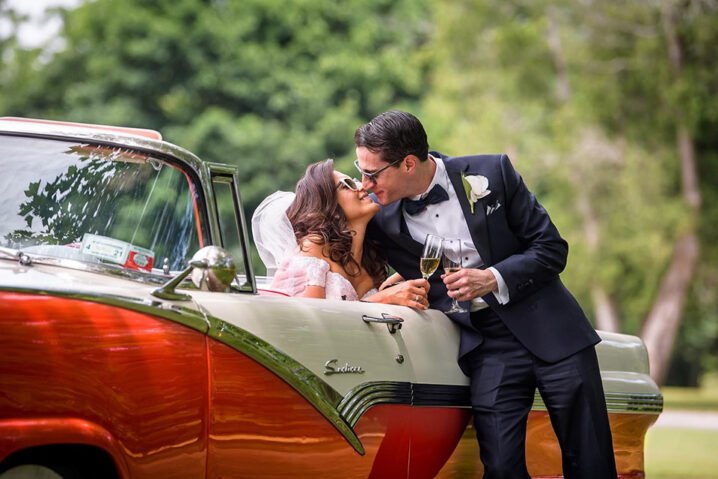 New York City's Premier Wedding Day Transportation Service 27 Bride and groom share a kiss while leaning on a classic vintage car, holding champagne glasses in a lush outdoor setting.