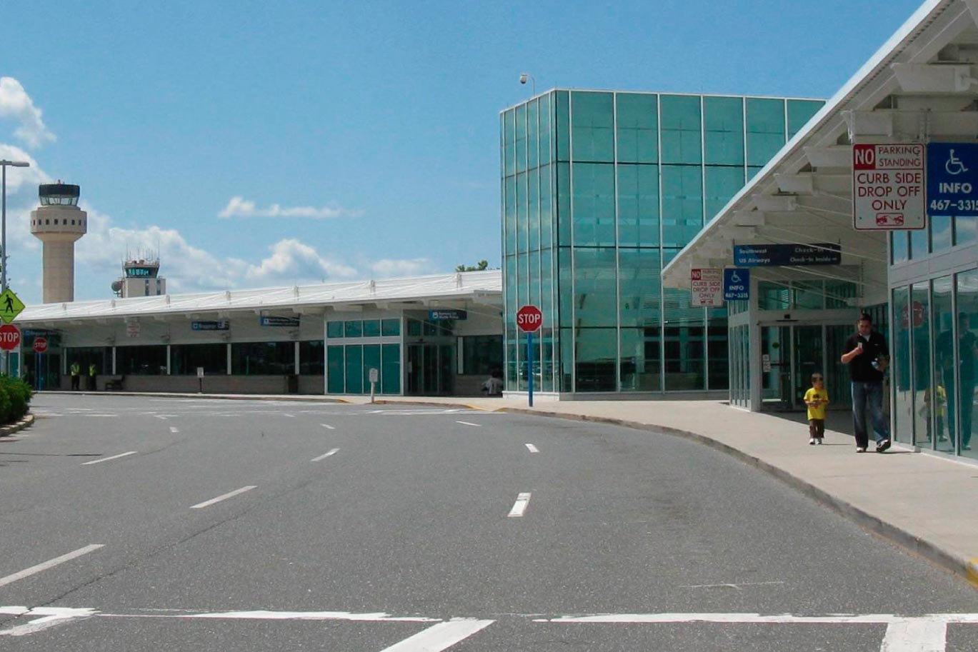 Street-level view of an airport terminal entrance with glass walls and a control tower in background.