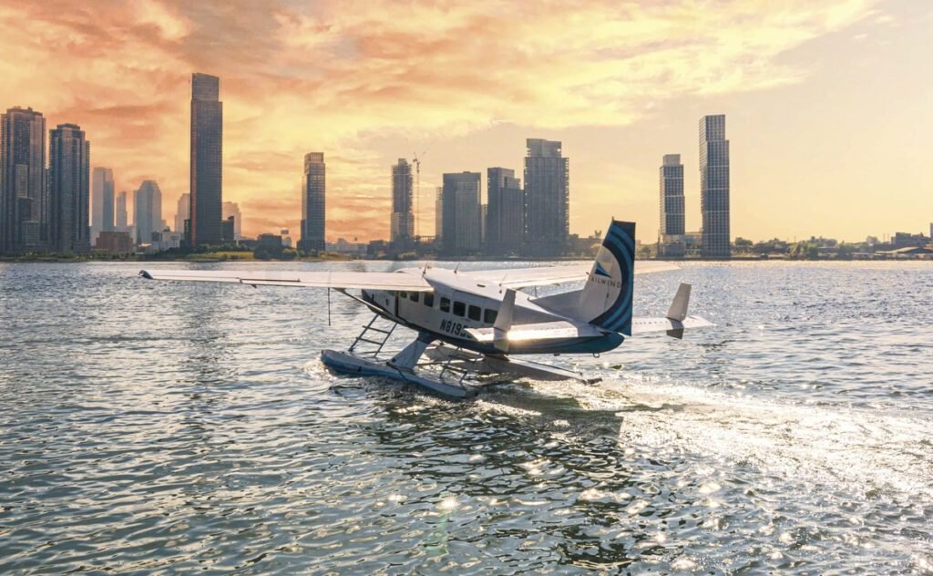 A seaplane taxiing on the water at sunset with a modern city skyline in the background, casting reflections on the surface.