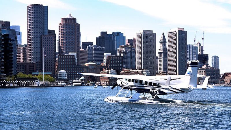 A seaplane taxiing on the water at sunset with a modern city skyline in the background, casting reflections on the surface.