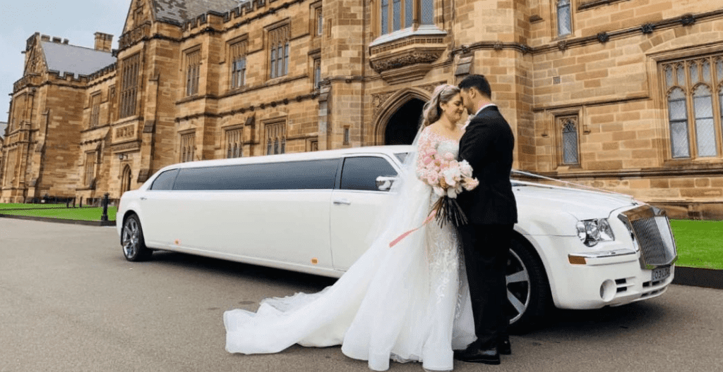 Bride and groom embracing in front of a white stretch limousine parked outside a historic sandstone building, ideal for wedding transportation.