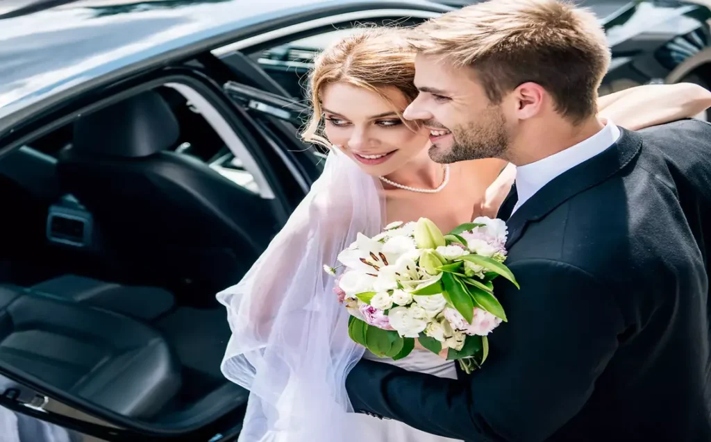Special Amenities and Services Offered for Wedding Transportation in New York 15 Happy bride and groom standing beside a luxury car, holding a bouquet of white flowers on their wedding day