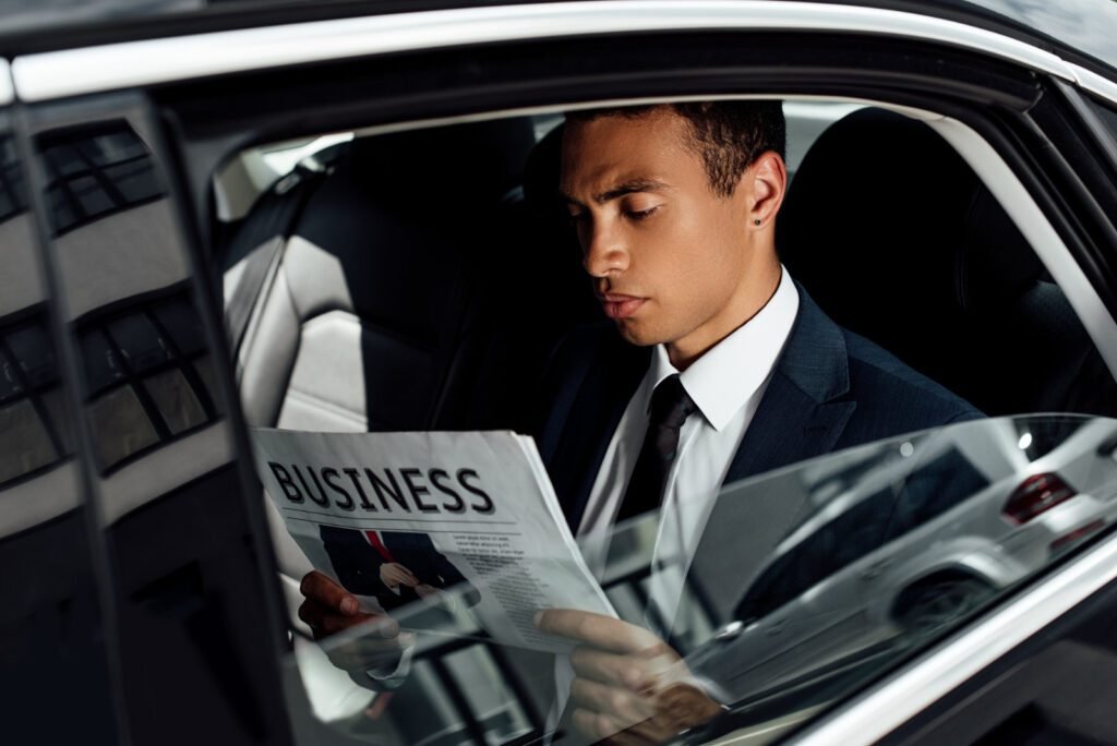 Businessman in a suit sitting in the backseat of a car, reading a business newspaper with a focused expression.