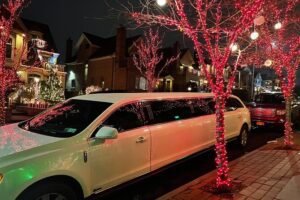 A white limousine is parked on a festive residential street at night, surrounded by trees wrapped in glowing red holiday lights.