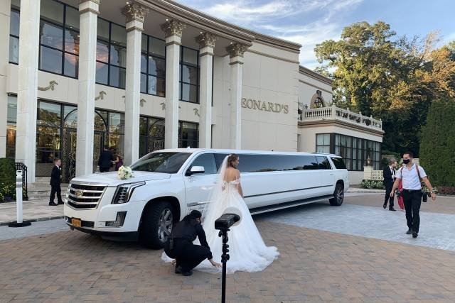Wedding Transportation Guide: Everything You Need to Know 18 Bride beside a white stretch limo at wedding venue, assistant fixing dress, photographer in background.