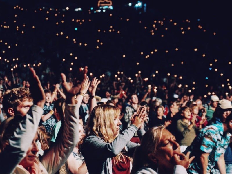 A cheering crowd at a lively concert, with people clapping and holding up phones in a dark venue filled with lights.