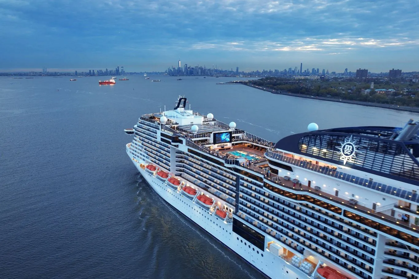 Large MSC cruise ship sailing near a city skyline at sunset with pools and deck lights visible.
