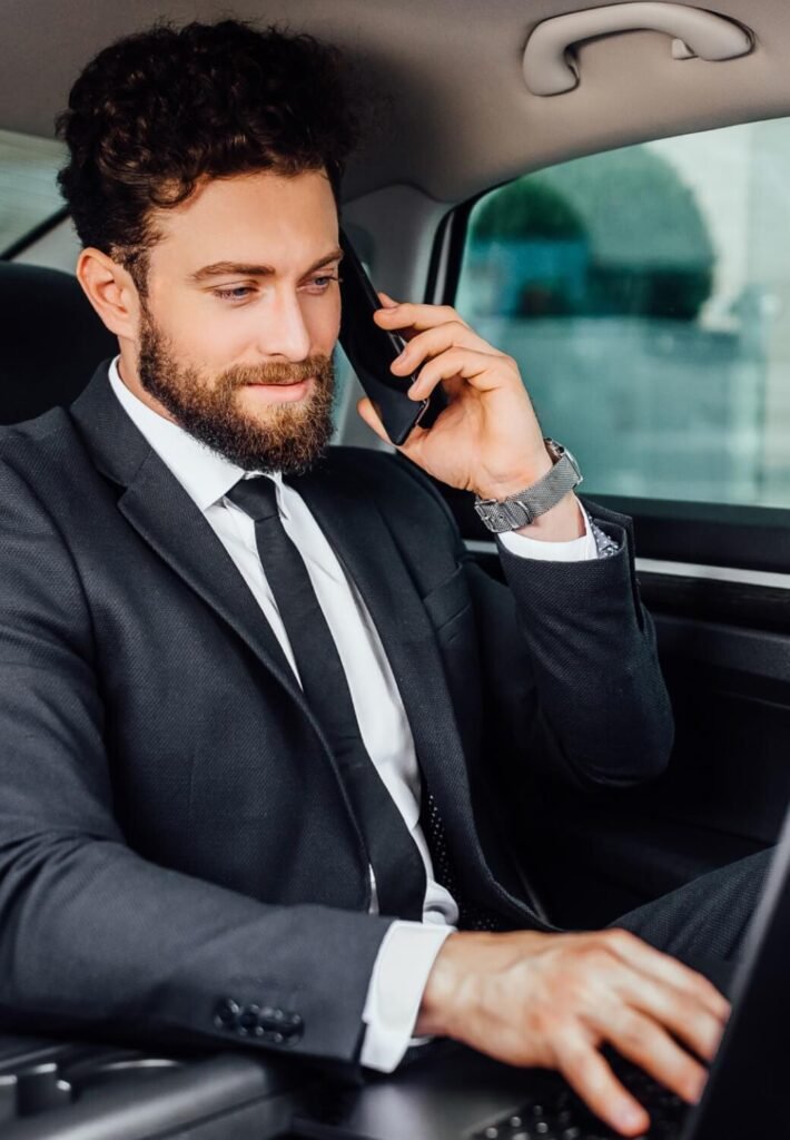 Business professional in a luxury car, speaking on the phone and working on a laptop, highlighting point-to-point car service.