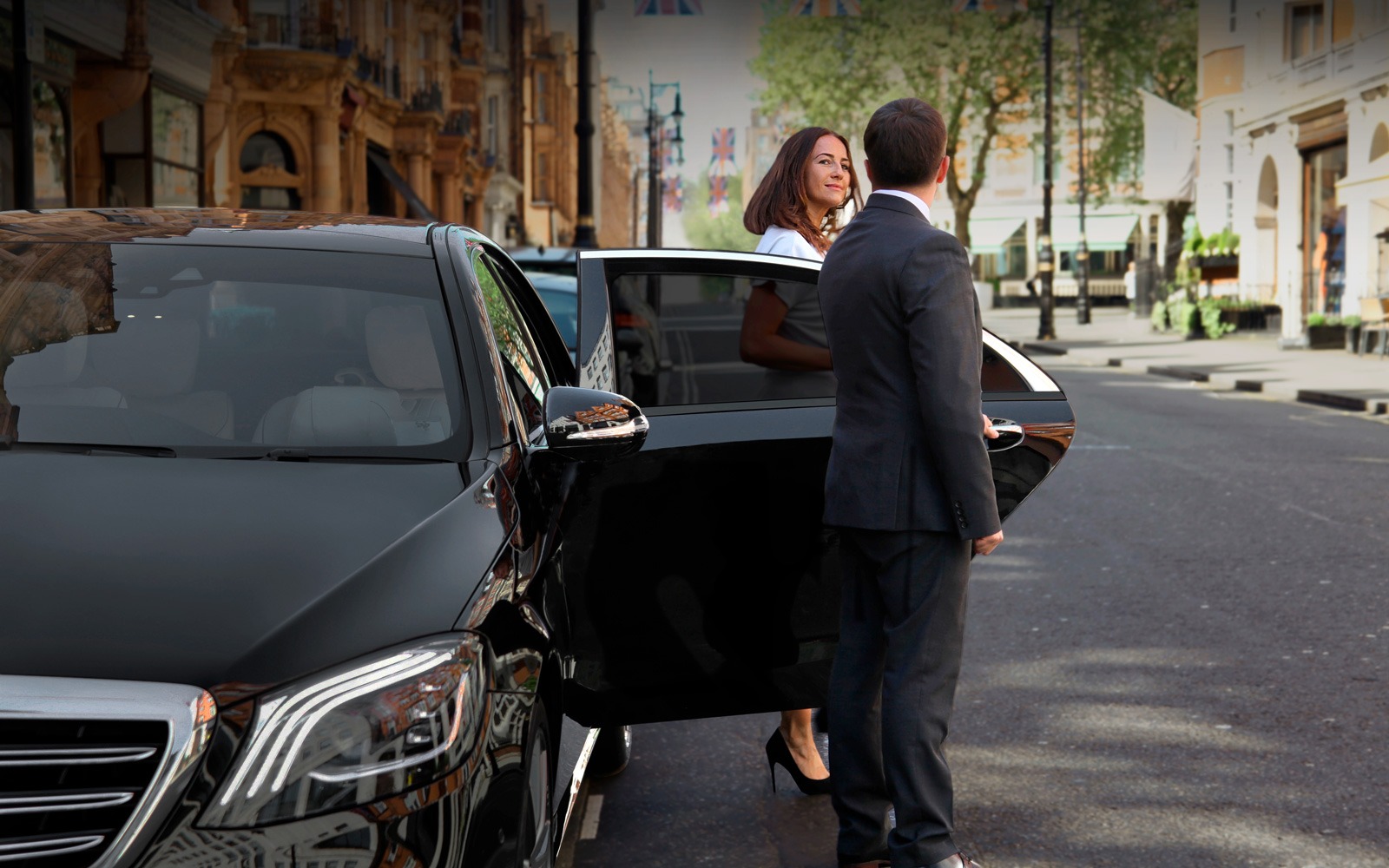 Chauffeur in a suit opening the door of a luxury black sedan for a woman in a city setting, symbolizing VIP chauffeur service.
