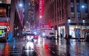 Rainy night in NYC with neon lights reflecting off wet streets, Radio City Music Hall glowing, traffic and pedestrians.