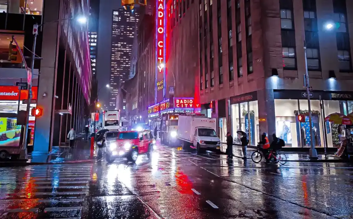 Rainy night in NYC with neon lights reflecting off wet streets, Radio City Music Hall glowing, traffic and pedestrians.