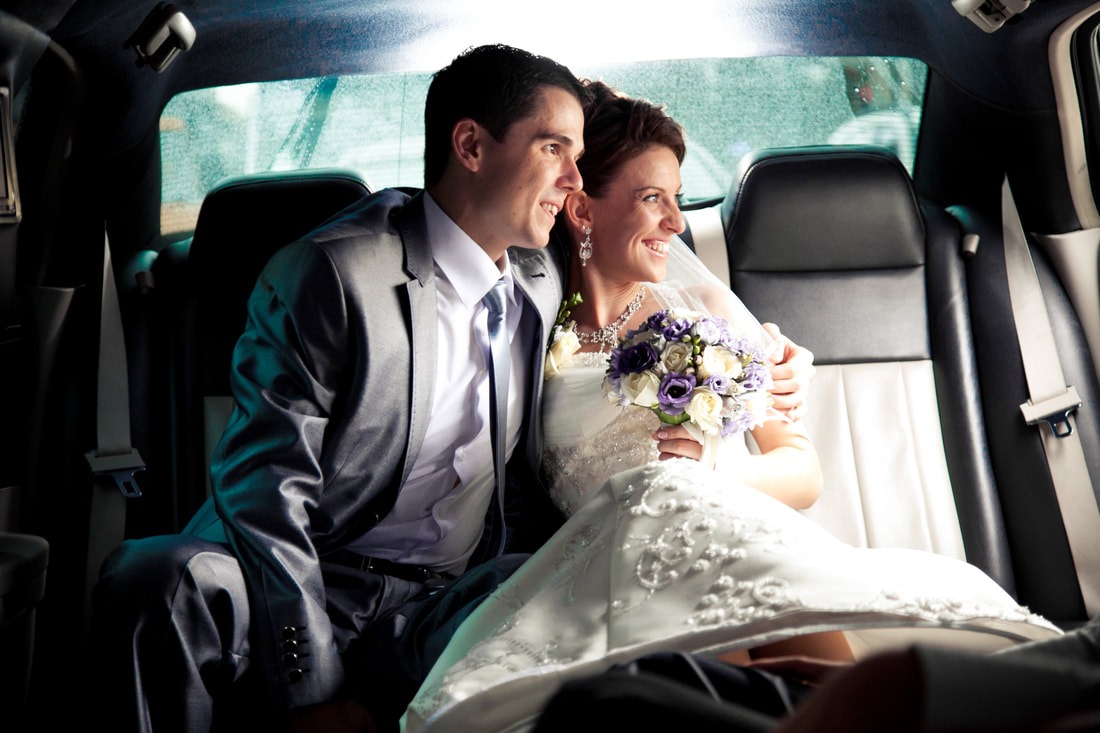 Happy newlywed couple sitting together in the backseat of a luxury limousine, smiling and holding a bouquet of purple and white flowers.