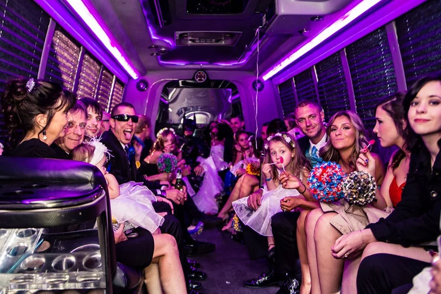 Party Limousine 2 Wedding party inside a limo bus with purple lights, guests smiling and holding flowers, kids and adults seated on both sides in celebration.