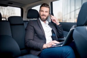 Businessman in a luxury car, working on a laptop and speaking on the phone during point-to-point car service ride.