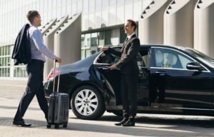 Chauffeur opening the door of a black private car for a businessman with luggage, standing outside a modern building."