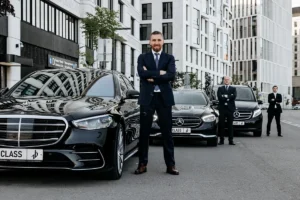 Professional chauffeurs in suits standing beside luxury black Mercedes-Benz vehicles in a modern city setting, representing premium limo service.